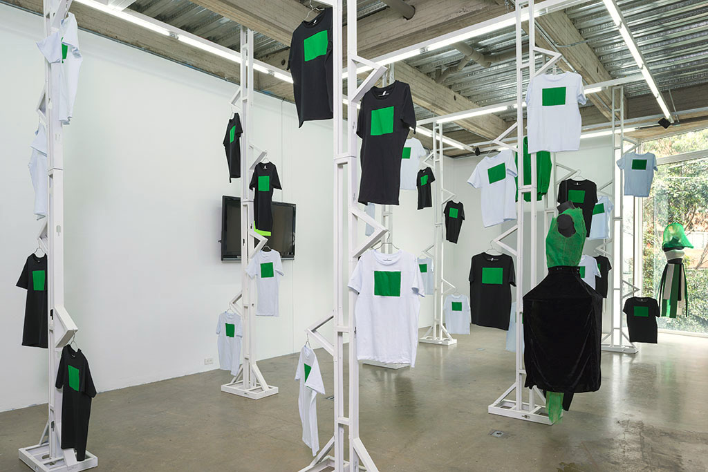 Exposición 'Primer Piso de la exposición Again_ en Espacio el Dorado, camisetas con pantalla verde colgadas alrededor de la sala.